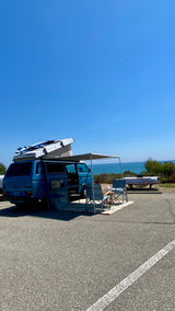 Scenic RV rental setup: blue VW Westfalia van with pop-top & surfboards by the ocean. Perfect for beach camping/van life.