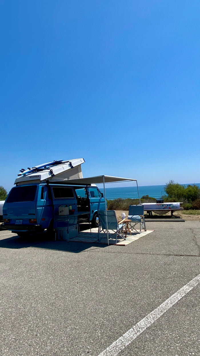 Scenic RV rental setup: blue VW Westfalia van with pop-top & surfboards by the ocean. Perfect for beach camping/van life.