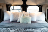 Inside a cozy RV rental: a neatly made bed with white pillows, a soft blanket, and a smiling llama plush.