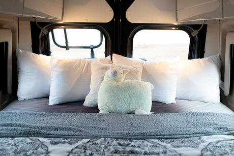 Inside a cozy RV rental: a neatly made bed with white pillows, a soft blanket, and a smiling llama plush.