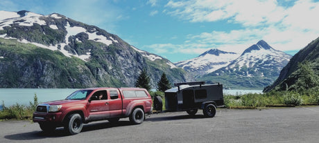 Explore stunning mountains with this red truck & adventure rv rental trailer by a glacial lake. Your next journey starts here!