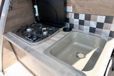 Inside an RV rental: A compact kitchenette with a two-burner FlameKing stove, sink, wood-grain counter, and checkered backsplash.