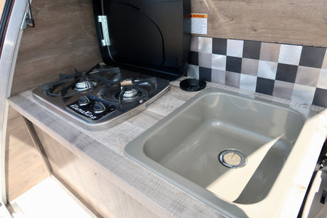 Inside an RV rental: A compact kitchenette with a two-burner FlameKing stove, sink, wood-grain counter, and checkered backsplash.