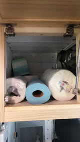 RV rental: Light wood overhead cabinet, hinges visible, contains three paper towel rolls (white & blue) and essentials for campervan living.