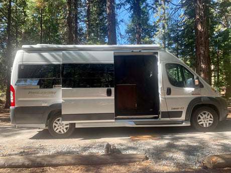 A light grey Pleasure-Way Lexor-TS RV with its side door open is parked at a sunny forest campsite. Great for an rv rental adventure!