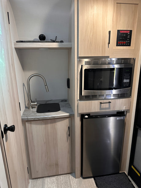 Compact RV rental kitchenette with a stainless steel microwave, mini-fridge, sink, and Winnebago control panel.