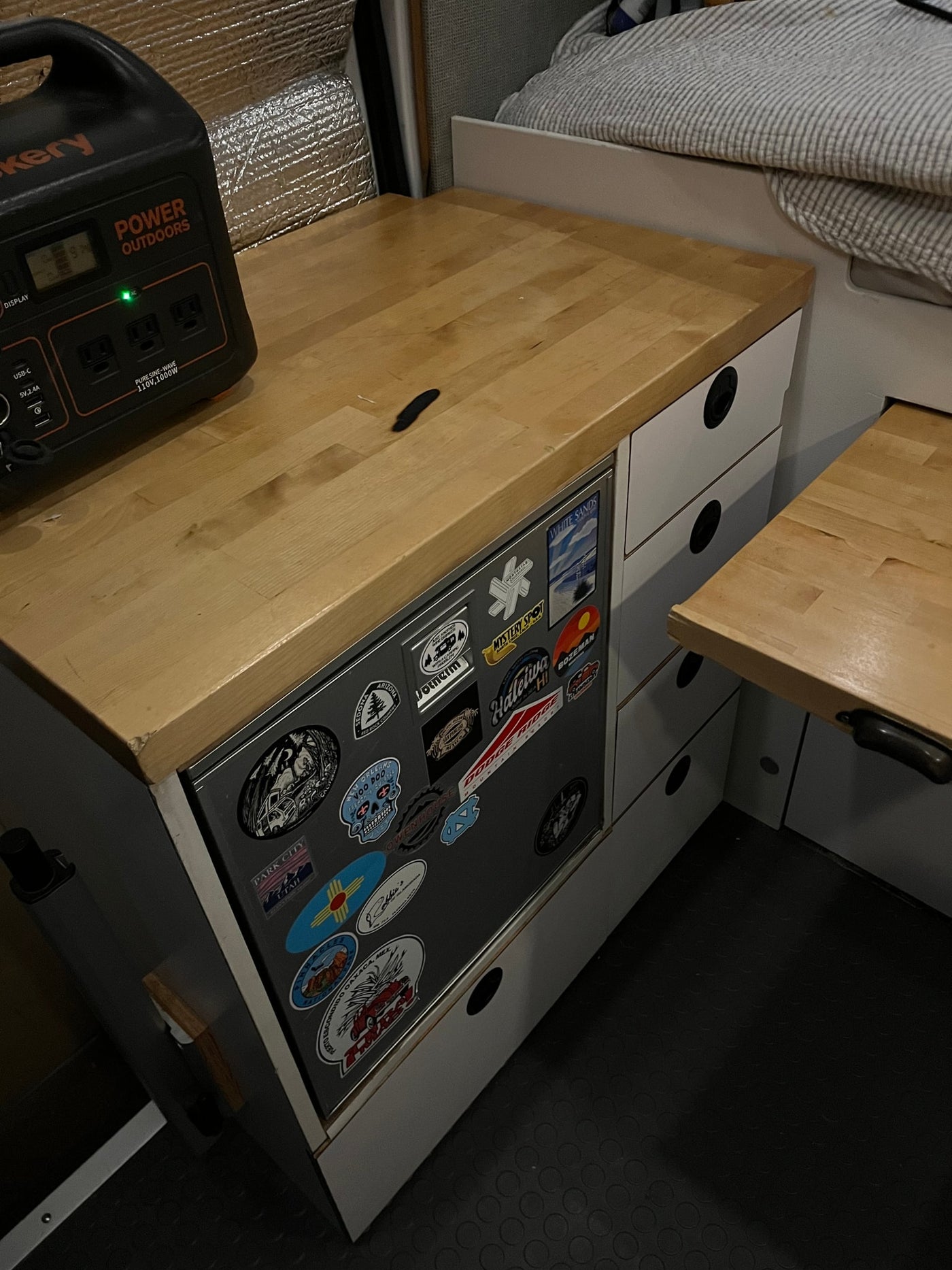 Inside an RV rental: A Jackery power station sits on a light wood counter with a sticker-covered fridge and storage drawers below. — photo 7