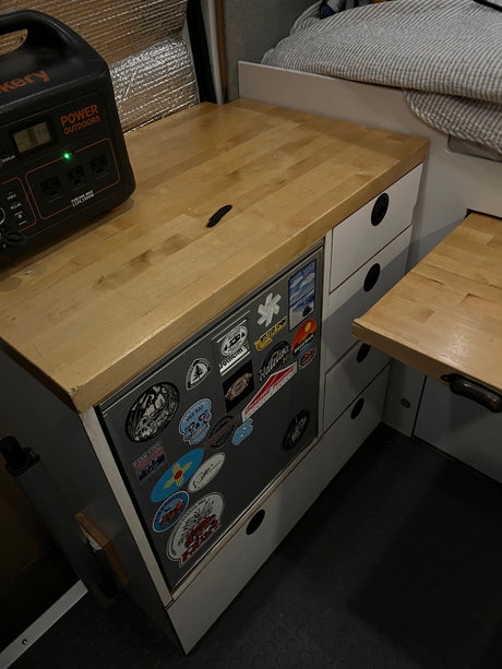 Inside an RV rental: A Jackery power station sits on a light wood counter with a sticker-covered fridge and storage drawers below.