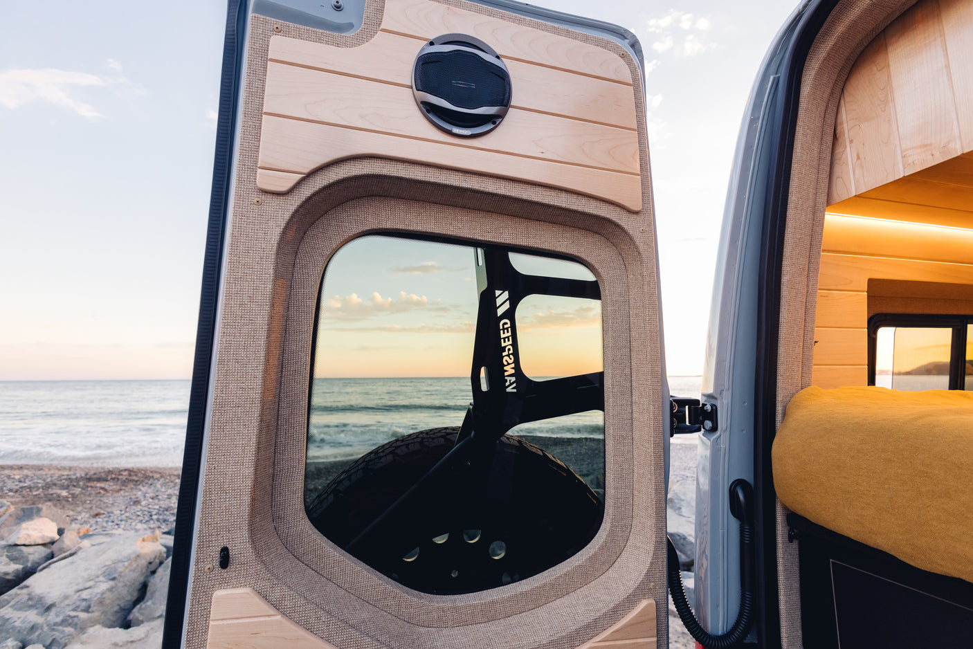 RV rental camper van interior view from open back door. Speaker, wood panels, bed inside, with ocean & sunset visible.