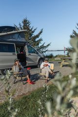 Two friends enjoy a sunny day next to their RV rental camper van, parked by the water for an outdoor adventure.