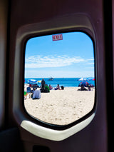 Beach vacation from an RV rental: sunny view of ocean, boats, people, and umbrellas through the window, with an EXIT sign.