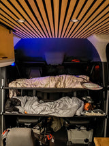 Inside an rv rental, a camper van's cozy interior features a slatted ceiling, blue accent lighting, and bunk beds with one person asleep.