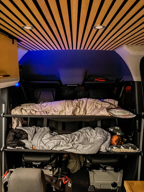 Inside an rv rental, a camper van's cozy interior features a slatted ceiling, blue accent lighting, and bunk beds with one person asleep.