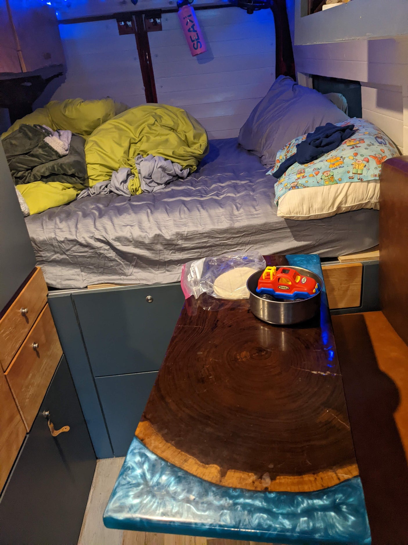 Inside a compact RV rental, a bed with colorful bedding, a custom epoxy table with a toy truck, and blue lights.