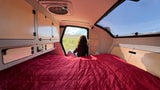Cozy RV rental interior: A person enjoys mountain views from the open hatch, with a comfy bed and storage.