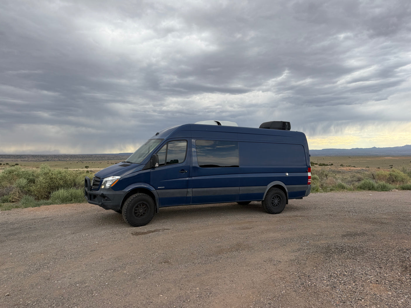 Dark blue Mercedes Sprinter camper van for rv rental on gravel road under stormy sky in arid landscape.
