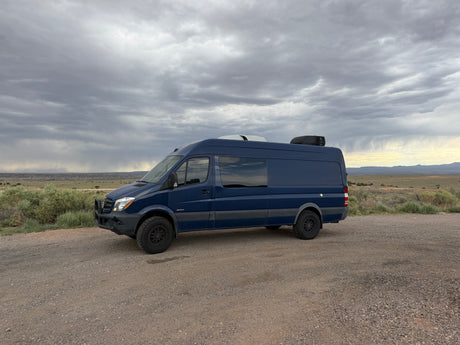 Dark blue Mercedes Sprinter camper van for rv rental on gravel road under stormy sky in arid landscape.