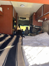 Cozy RV rental interior with a comfortable bed, overhead cabinets, and the driver's area visible ahead.