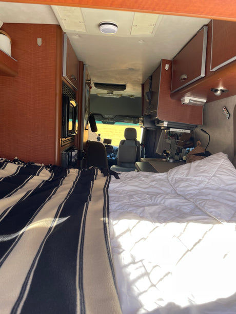 Cozy RV rental interior with a comfortable bed, overhead cabinets, and the driver's area visible ahead.