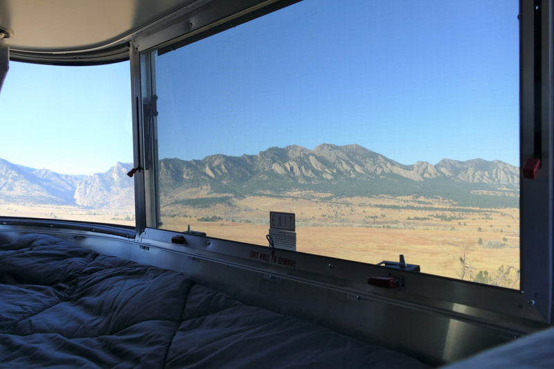An RV rental's cozy bed and window with a screen showcasing rugged mountains and a golden prairie under a clear sky. — photo 19