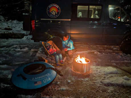 An RV rental provides cozy winter camping. Two people warm up by a bright campfire next to their van in the snow. Zangeri tube nearby.