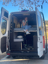 Custom camper van RV rental interior: a woman relaxes on the bed with skateboards stored below.