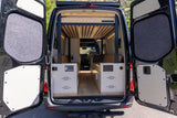 Discover the cozy interior of this custom RV rental van. Open rear doors reveal a built-in bed, control panel, and storage.