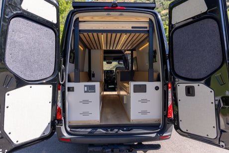 Discover the cozy interior of this custom RV rental van. Open rear doors reveal a built-in bed, control panel, and storage.
