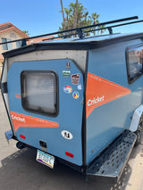 A light blue Cricket camper, ready for adventure. This RV rental sports a roof rack and travel stickers.