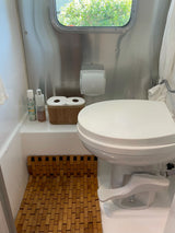 RV rental bathroom with a white foot-flush toilet, shiny stainless walls, a bamboo mat, and toiletries.