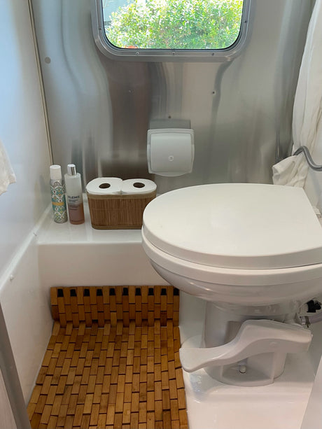 RV rental bathroom with a white foot-flush toilet, shiny stainless walls, a bamboo mat, and toiletries.