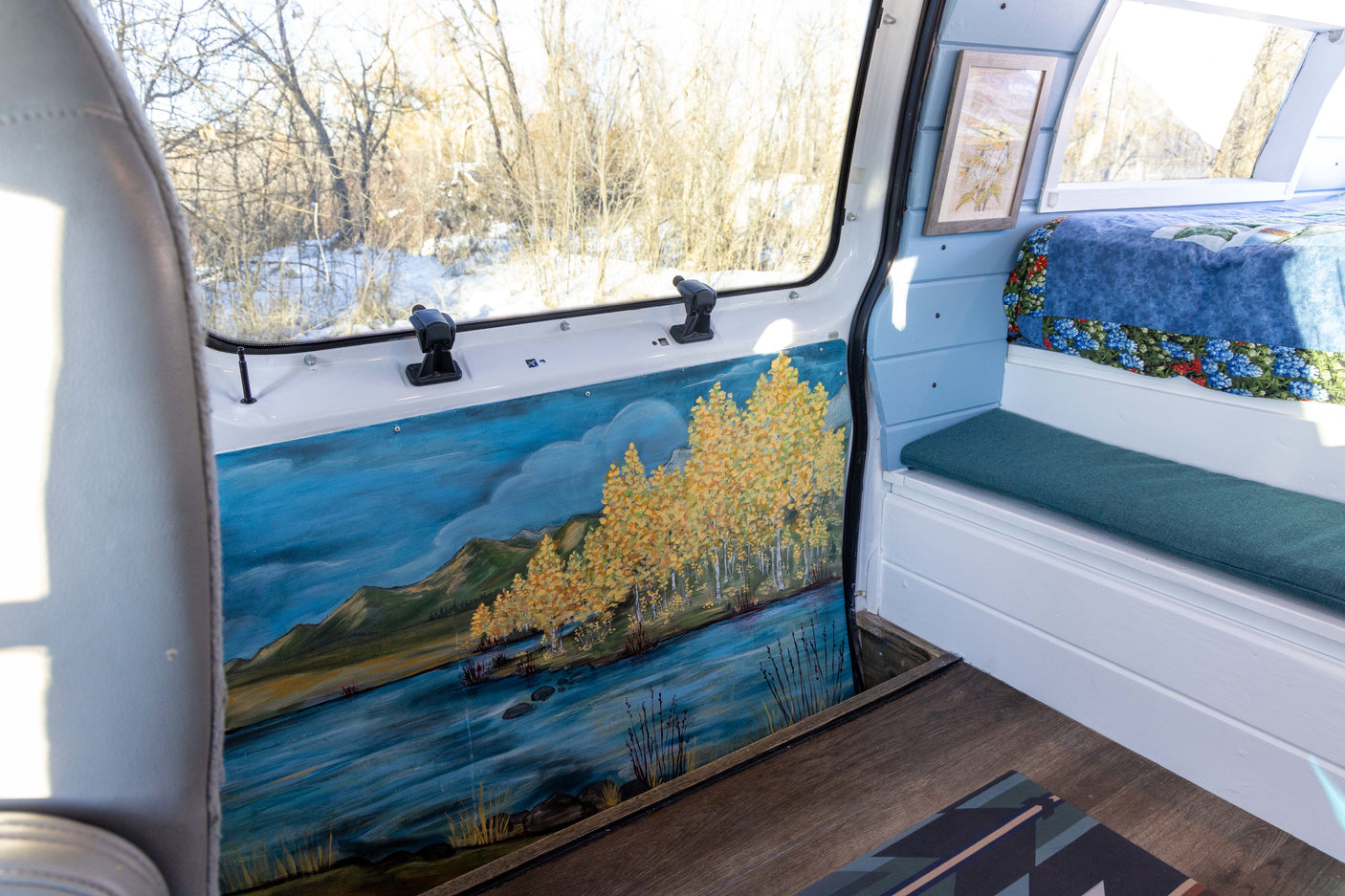 This RV rental's custom campervan interior features a scenic painted wall, cozy bench, bed, and snowy window view.