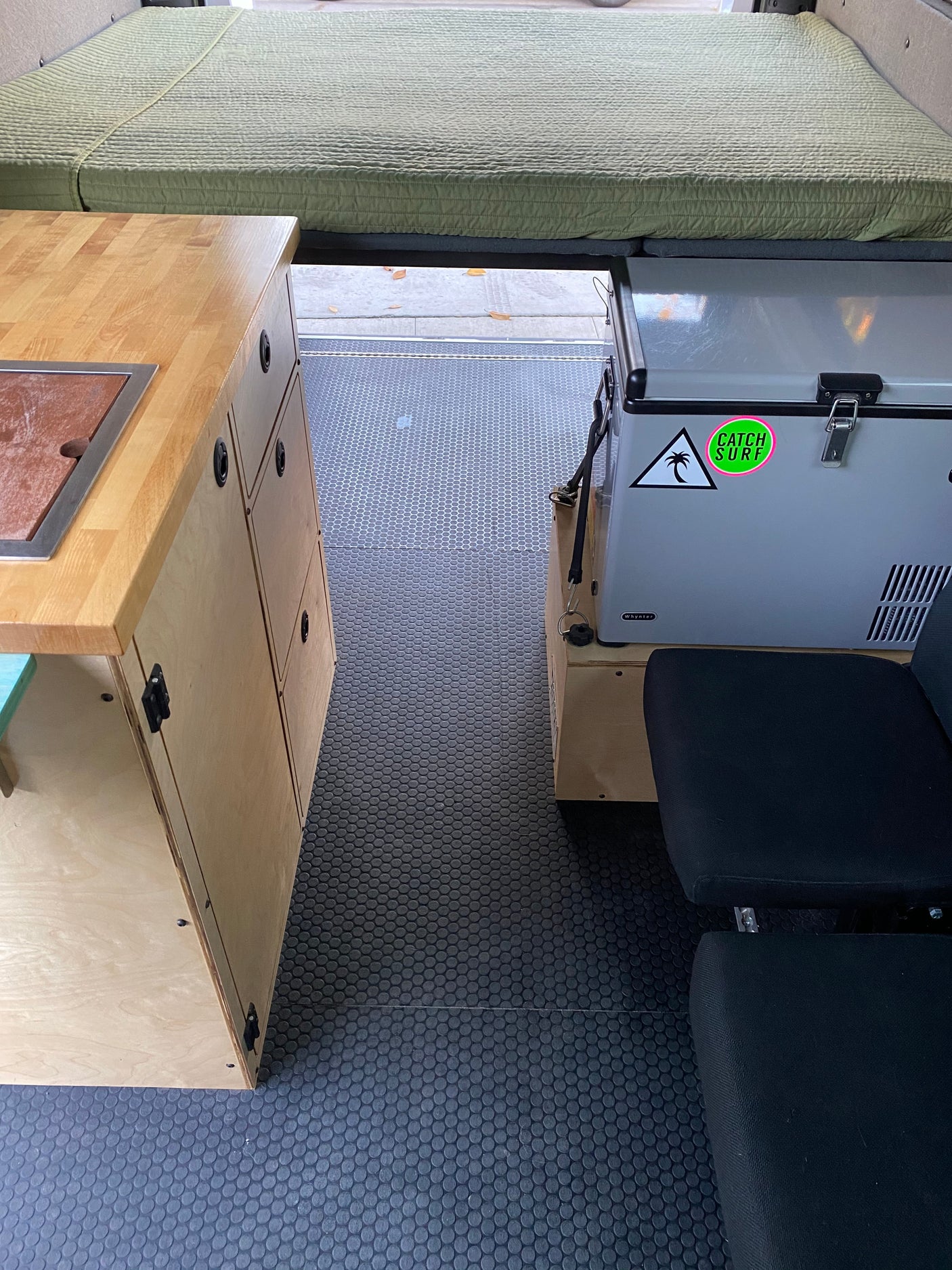 RV rental interior shows a green bed, wood kitchen counter, and portable fridge next to a black seat. Dark patterned floor.
