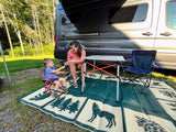 Mother & child relax on a mat by their rv rental camper van at a sunny campsite.
