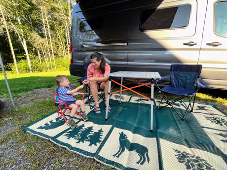 Mother & child relax on a mat by their rv rental camper van at a sunny campsite.