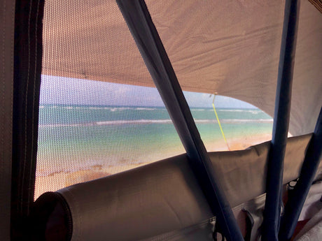 Ocean view from an RV rental through the mesh screen, showing a sandy beach and blue-green water.