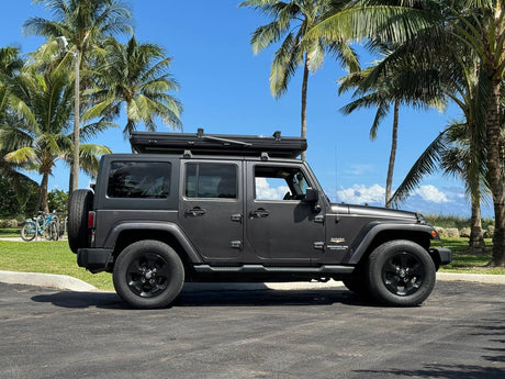 This dark grey Jeep Wrangler, equipped with a rooftop tent for RV rental adventures, is parked under palm trees on a sunny day.