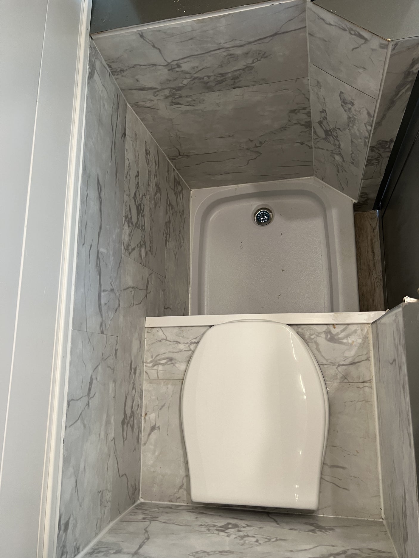Compact RV rental bathroom with a white toilet and corner shower, featuring grey marble-look walls and floor.