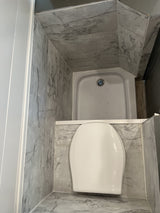 Compact RV rental bathroom with a white toilet and corner shower, featuring grey marble-look walls and floor.