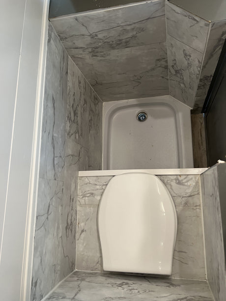 Compact RV rental bathroom with a white toilet and corner shower, featuring grey marble-look walls and floor.