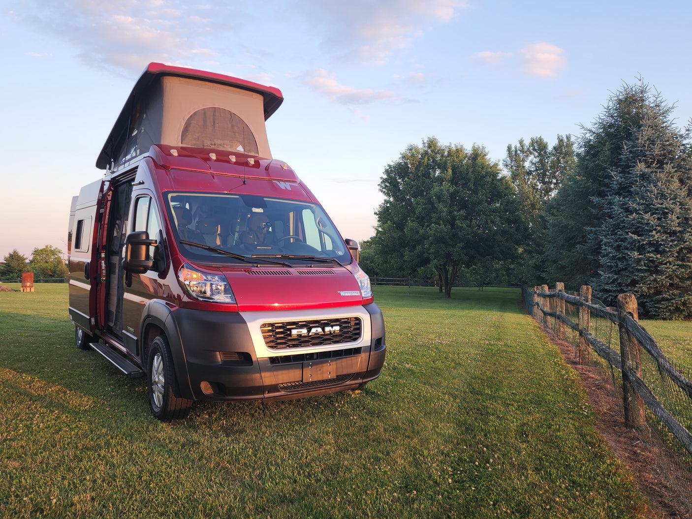 Red Winnebago camper van RV rental, pop-top open, ready for adventure on a grassy field at golden hour.