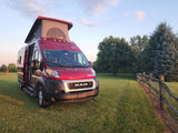 Red Winnebago camper van RV rental, pop-top open, ready for adventure on a grassy field at golden hour.