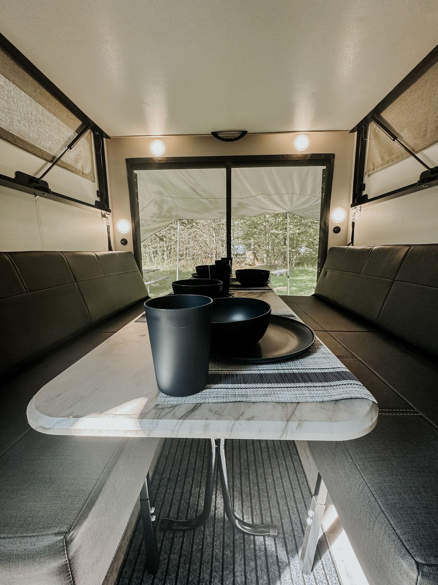 RV rental interior shows a set dining table with black dishes, surrounded by comfy benches, overlooking a forest.