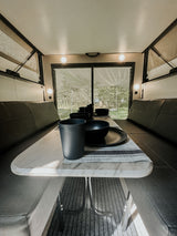 RV rental interior shows a set dining table with black dishes, surrounded by comfy benches, overlooking a forest.