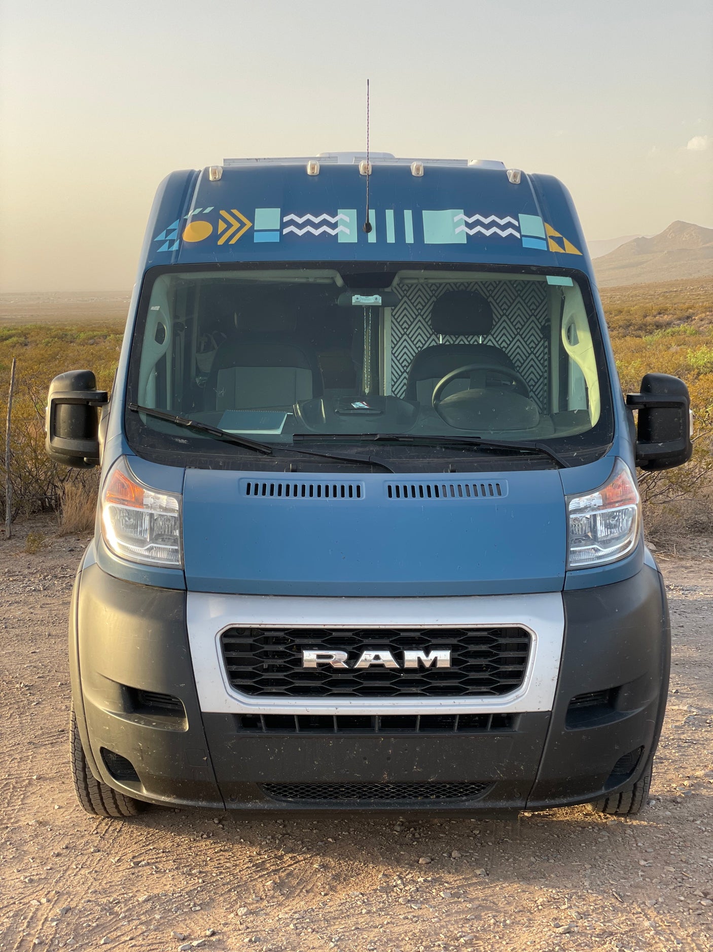 Front view of a blue Ram ProMaster campervan, an RV rental, parked on a dirt road in a desert setting.