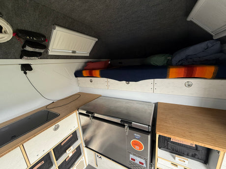RV rental: Compact interior featuring a bed with organized storage, a kitchenette with stove, and a portable fridge.