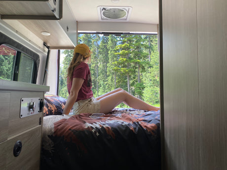 Enjoying nature from an RV rental, a woman relaxes on the bed, looking out at a lush forest view through the back window.