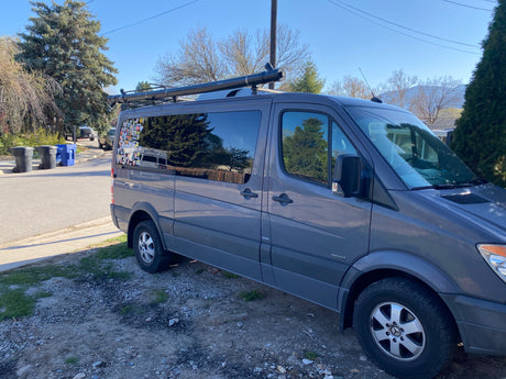 Gray Mercedes Sprinter van, an RV rental, with roof rack and rear window stickers, parked on gravel.