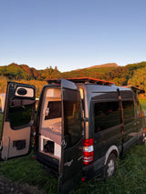 A dark Mercedes-Benz Sprinter rv rental camper van with open doors, revealing its bed, parked at a scenic campsite with mountains in golden hour light.