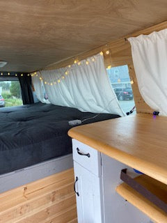 Interior view of an RV rental: wooden walls, string lights, a dark bed, and a light counter create a cozy camper van space.